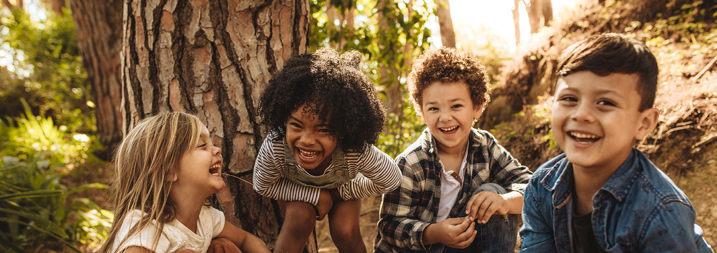 Vier Kinder im Kindergartenalter mit unterschiedlicher Hautfarben hocken im Wald und lachen in die Kamera