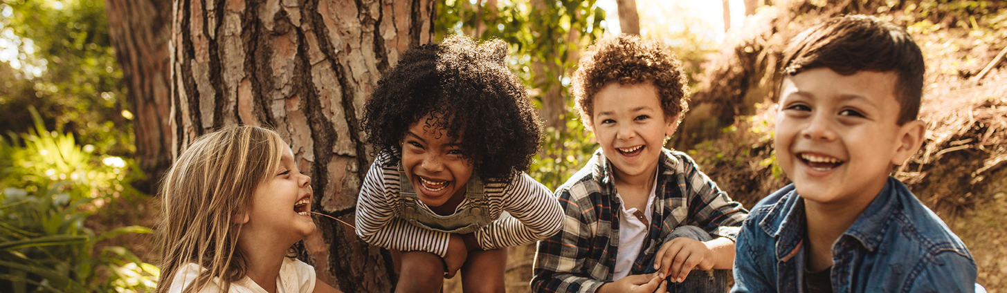 Vier Kinder im Kindergartenalter mit unterschiedlicher Hautfarben hocken im Wald und lachen in die Kamera