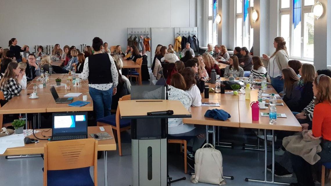 Im Konferenzraum sitzen viele Mitarbeiter an Tischen und verfolgen den Vortrag